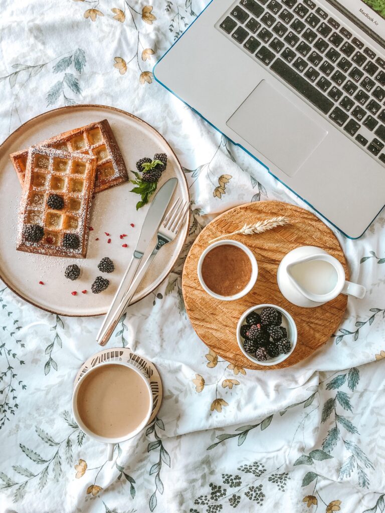 A laptop, a cup of coffee, and a plate with waffles and sauce are placed on a white tablecloth.