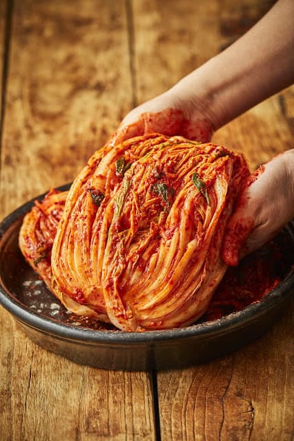 Hands holding red napa cabbage kimchi on top of a clay jar lid